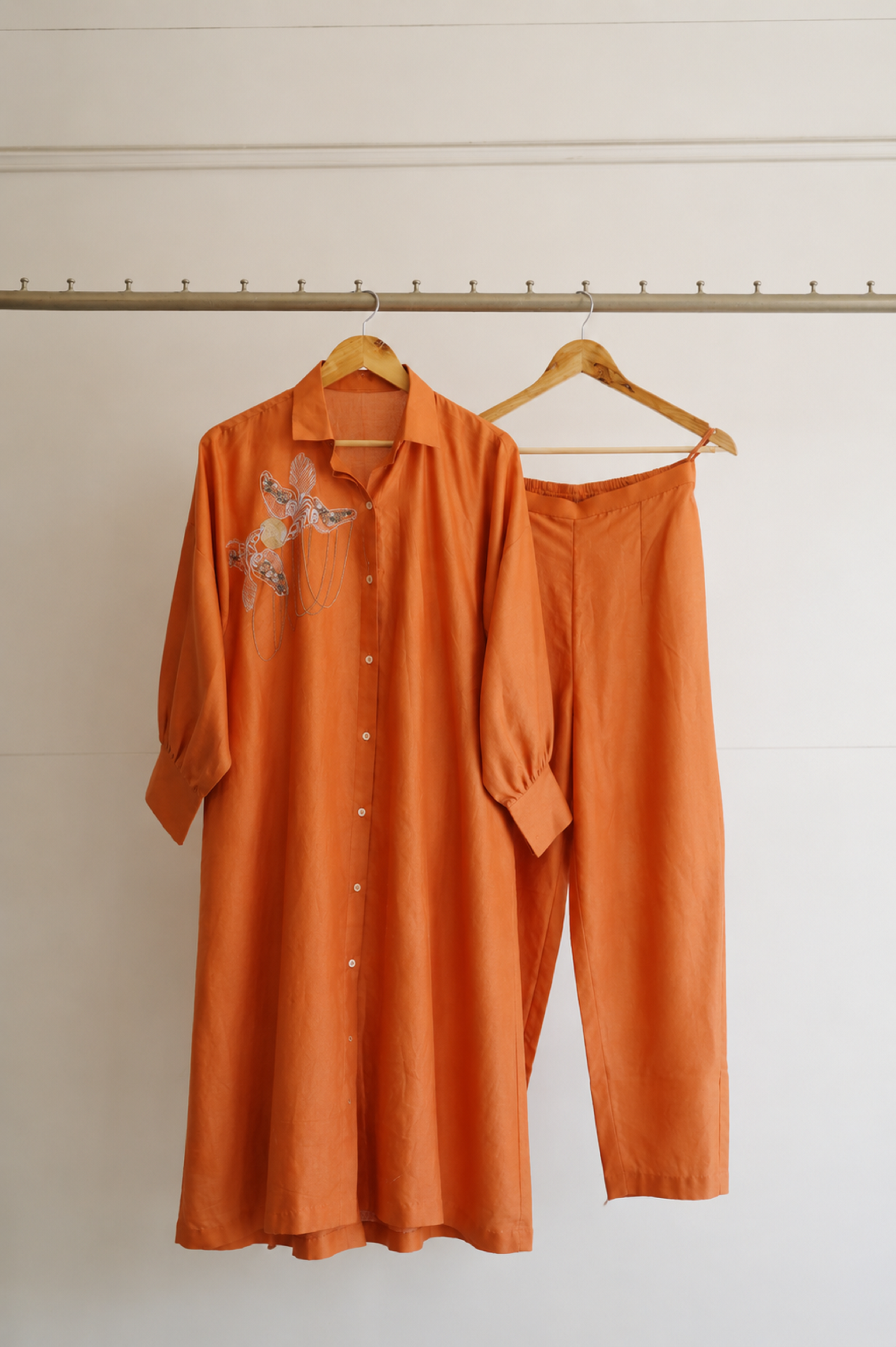 Tinka collection rust chanderi 3-piece set by eeda — long embroidered shirt with handwork. straight trousers and bustier, handcrafted in Varanasi, hung on wooden hangers against a light background